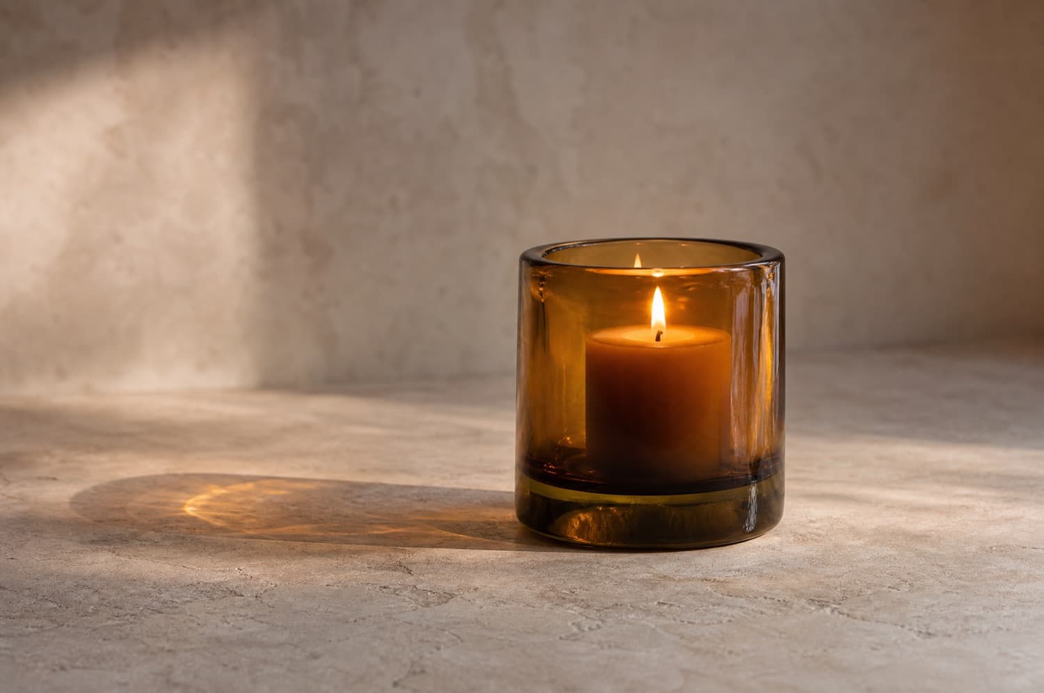 A thick amber glass candle holder with a wide pillar candle burning in it on a bare stone surface, well clear of anything else.