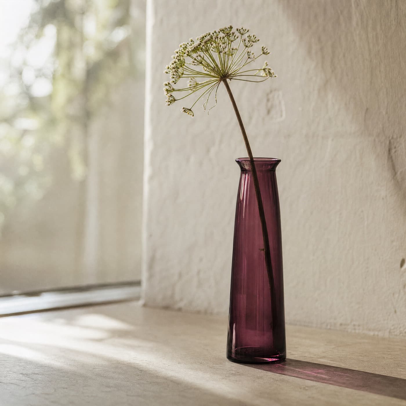 Narrow plum glass bud vase holding a single stem on a windowsill against bright light.