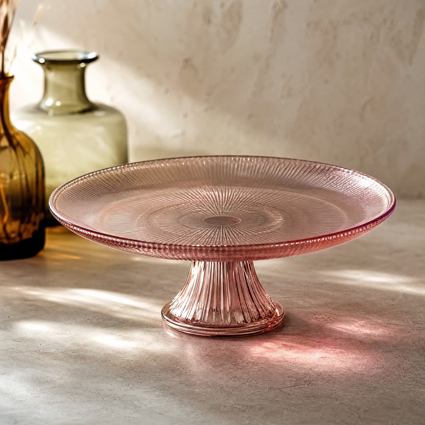 Footed misty rose glass cake plate on a table, a soft pink pool of light gathered beneath the foot.