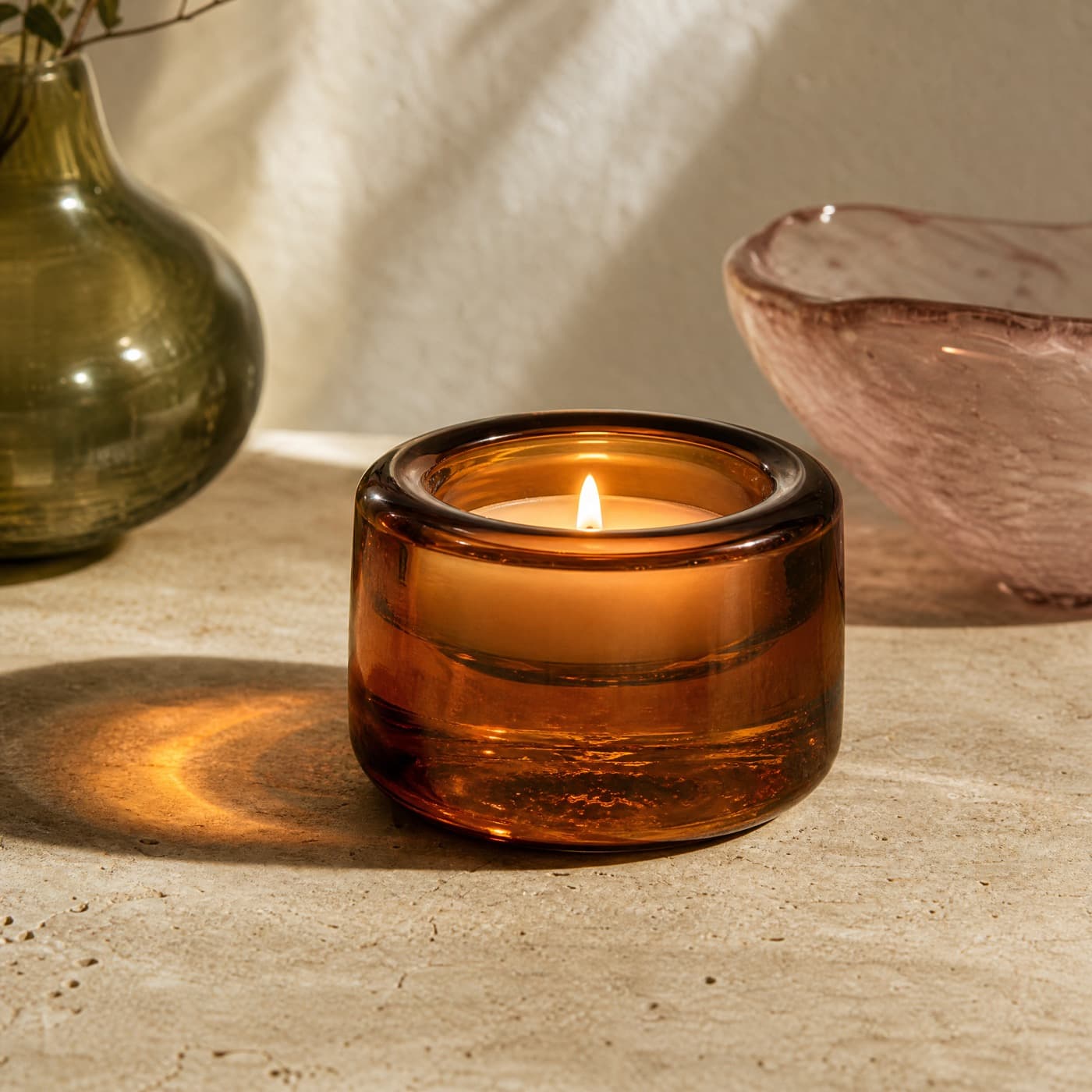 Heavy amber glass candle holder with a lit candle, warm light spilling outward across a dark table.