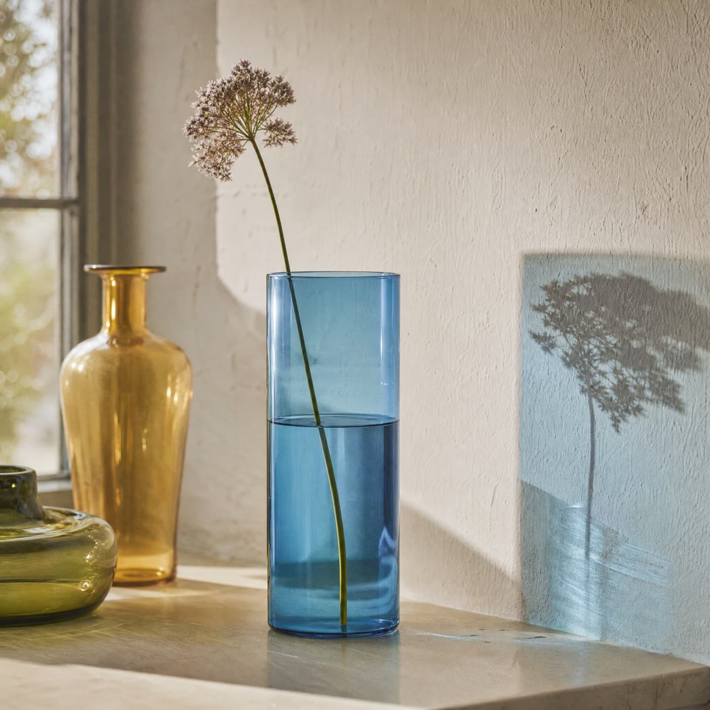 Sea-blue cylinder vase on a windowsill, projecting a tall blue rectangle of light onto the wall behind.