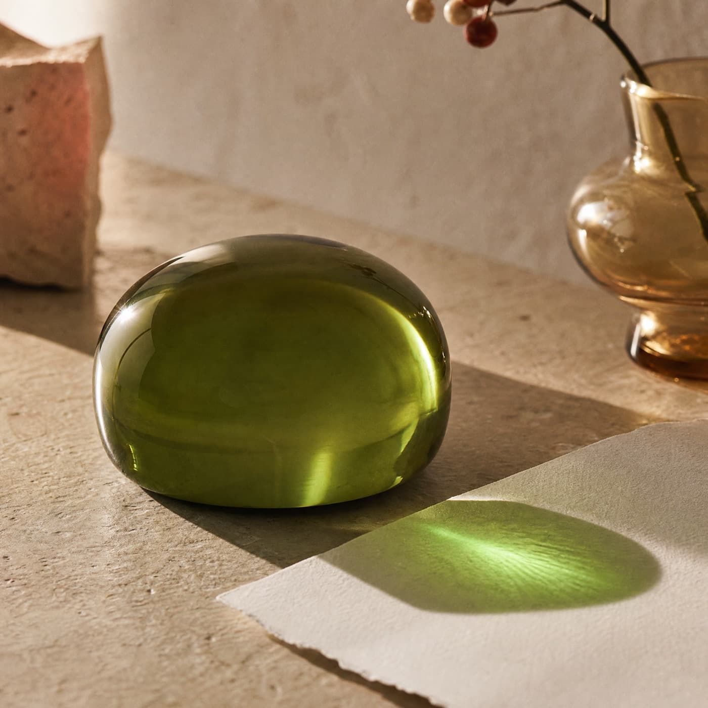 Solid glass-green paperweight on a desk in raking sun, casting a small deeply saturated green mark on paper.