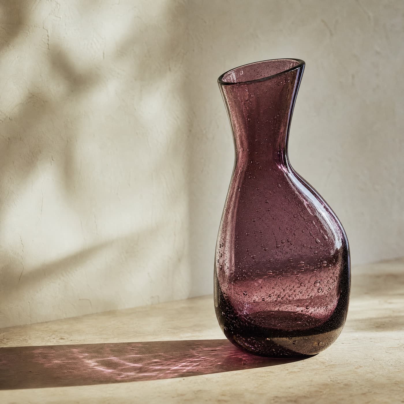Mouth-blown plum carafe with a leaning body and visible bubbles, strongly backlit.