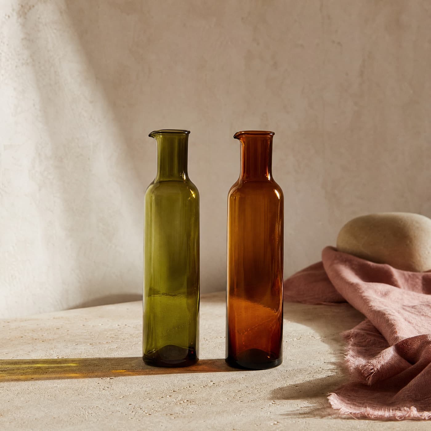 A green and an amber glass bottle with drawn pouring lips, side by side in raking window light.