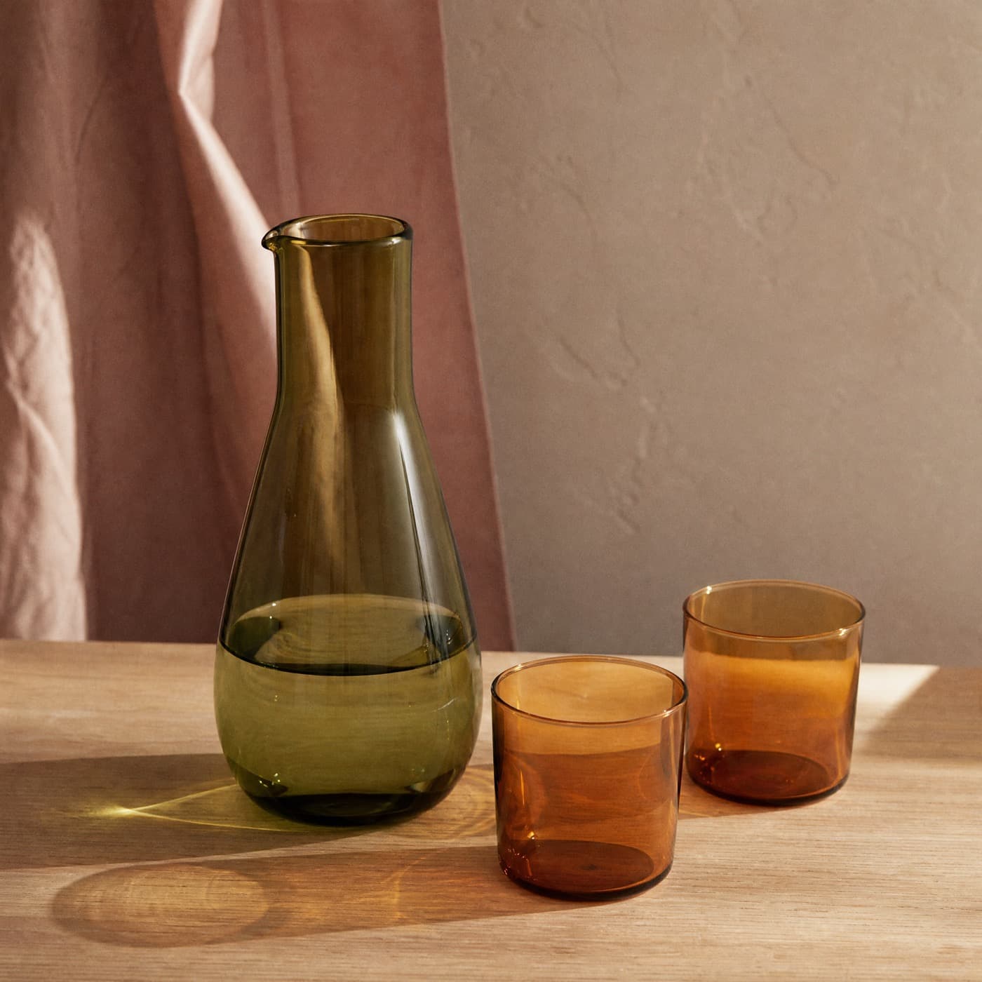 A green glass carafe with two amber glasses on an oak table, their coloured casts overlapping between them.