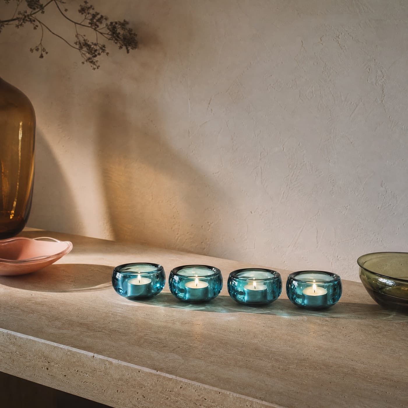 Four sea-blue glass tealight holders in a row on a dark table, their pools of blue light overlapping.