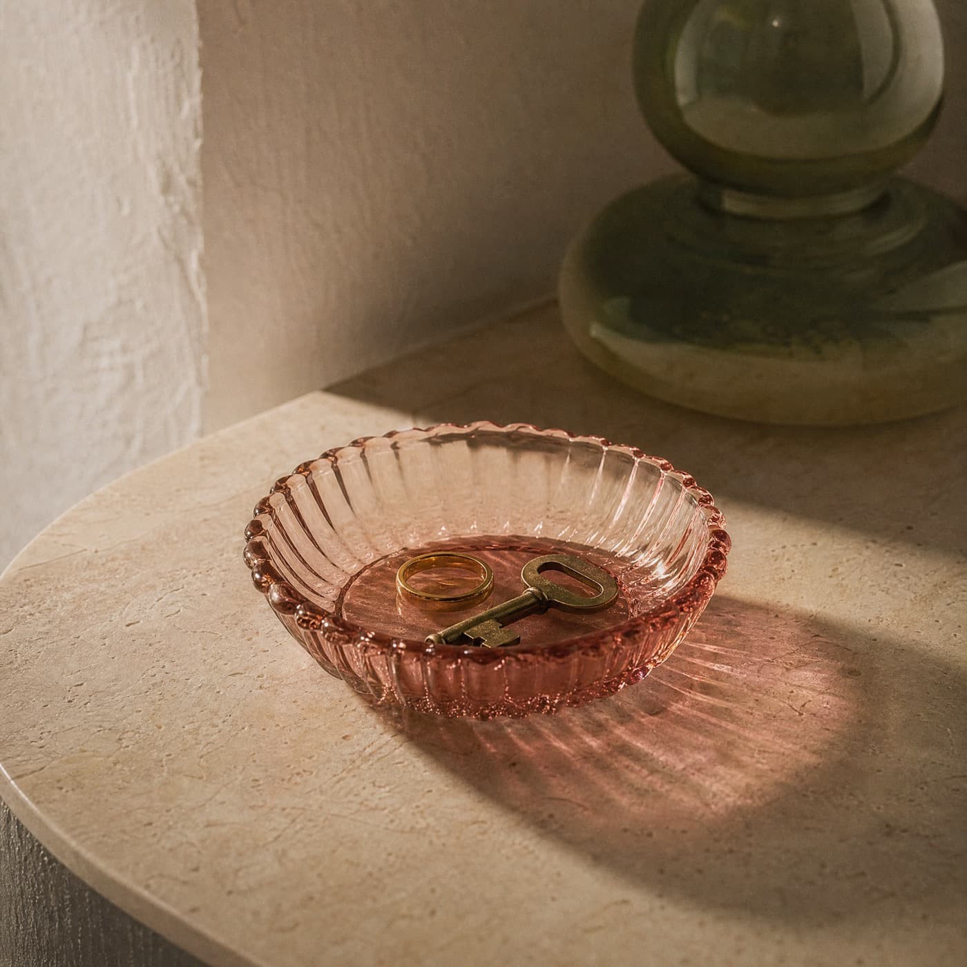 Shallow misty rose glass dish holding a ring and a key on a bedside table in low light.