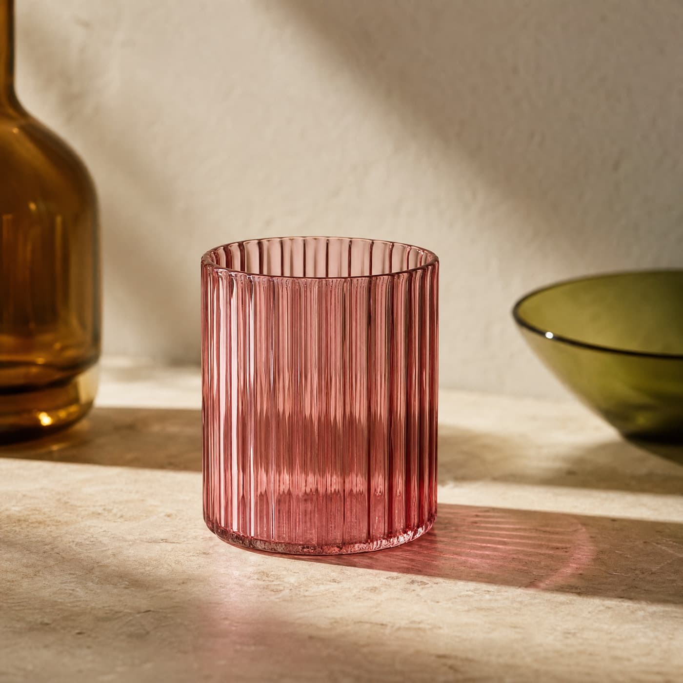 Ribbed misty rose tumbler in side light, throwing a striped pink patch onto a pale surface.