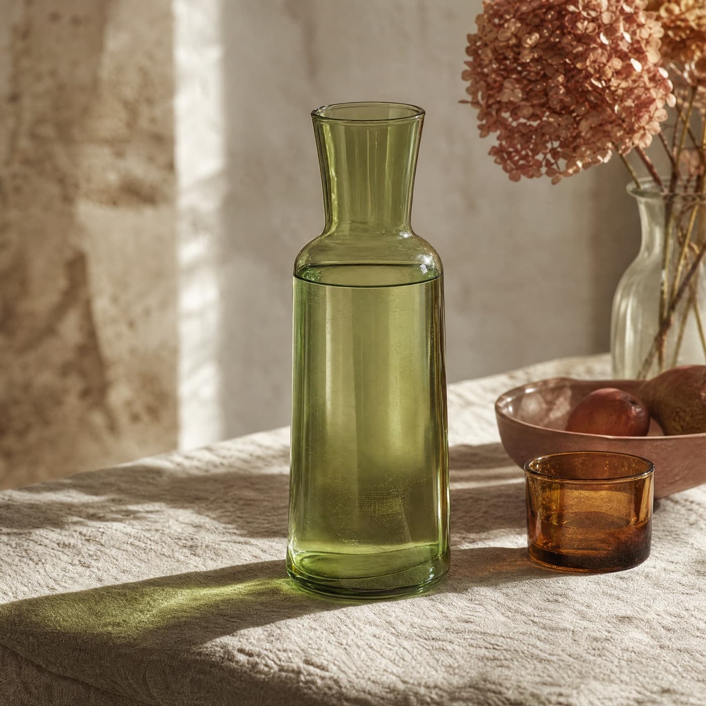 Glass-green one litre carafe filled with water, backlit, throwing a broad green patch across a linen tablecloth.