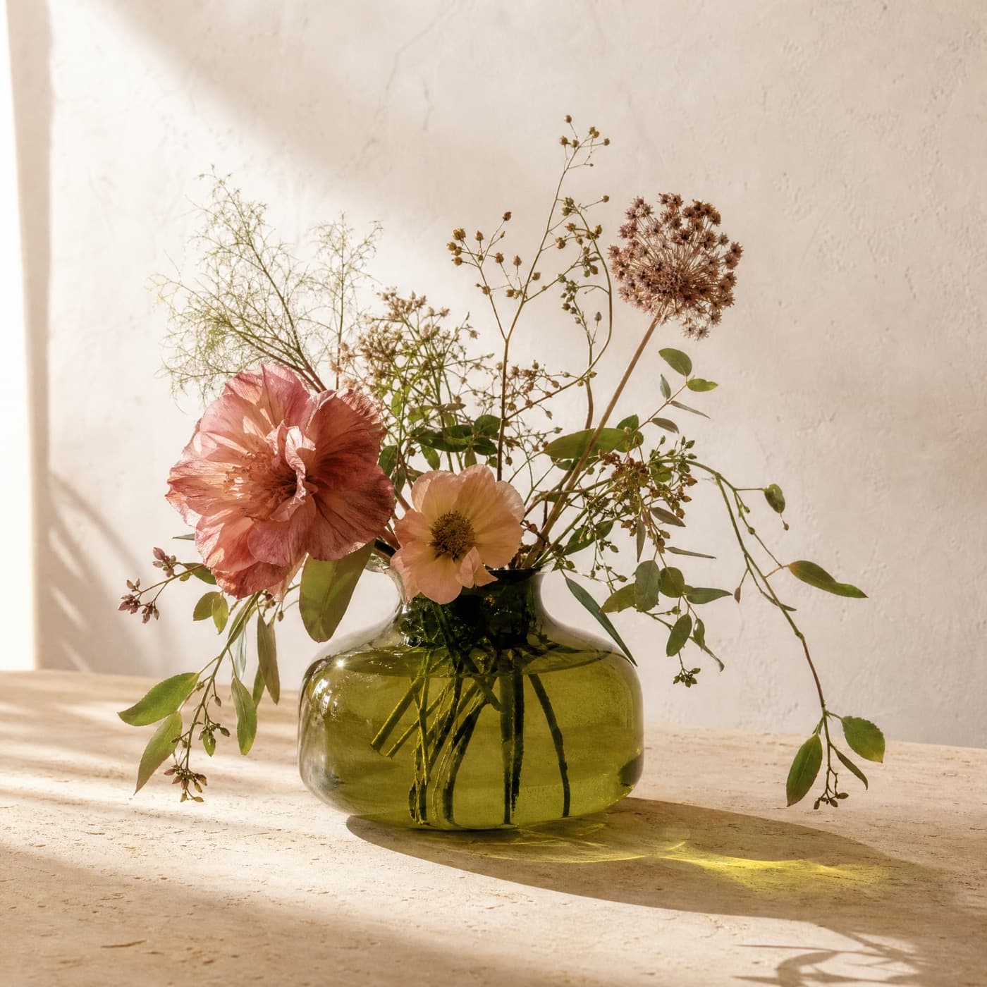 Low wide glass-green vase with stems falling outward, sunlight passing through onto a pale wall.