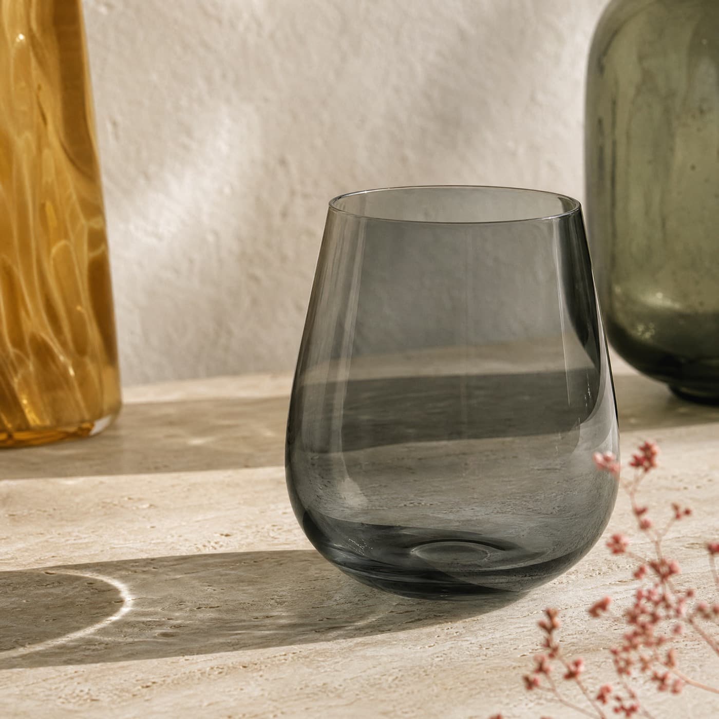 Smoke-grey stemless wine glass backlit on a pale oak table with a soft grey shadow of colour beneath it.